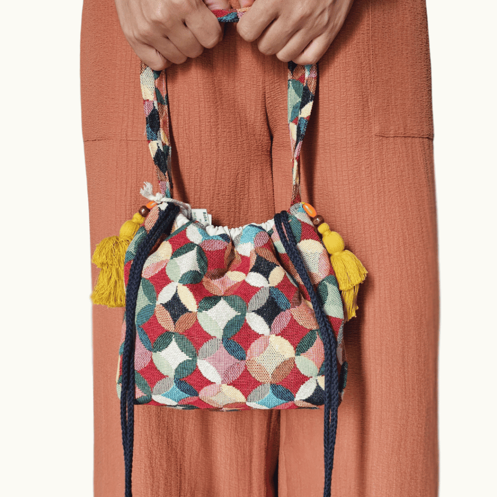 A stylish drawstring bucket bag or pouch bag featuring a vibrant, multi-color geometric fabric pattern with interlocking circle or Seigaiha wave motifs with bohemian style. The textile includes shades of teal, burgundy, olive green, cream, and brown. Decorated with tassels and wood beats