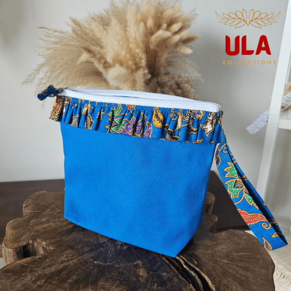 A vibrant zippered pouch bag or clutch with a solid royal blue cotton body. The bag is decorated with a wide, ruffled trim along the white zipper, made from a blue batik fabric with a multicolor floral print in shades of gold, purple, and red. The bag is shown open and includes a matching batik fabric wrist strap. It rests on a dark, rustic wooden surface, with a logo and dried pampas grass in the background, showcasing an eclectic fashion accessory perfect for travel or casual use.