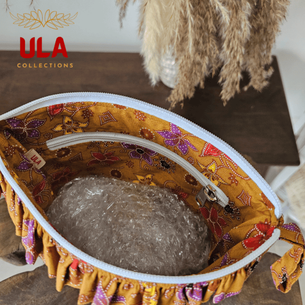 A stylish zippered pouch or clutch bag featuring a deep ochre brown cotton body with a subtle texture. The top is decorated with a wide, ruffled trim and interior lining made from a vibrant gold batik fabric with purple and brown floral motifs. This versatile fashion accessory includes a matching batik wrist strap and is shown styled as a handbag and resting on a wooden surface, offering an eclectic and bohemian accent for everyday or travel use.