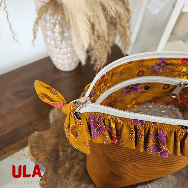 A stylish zippered pouch or clutch bag featuring a deep ochre brown cotton body with a subtle texture. The top is decorated with a wide, ruffled trim and interior lining made from a vibrant gold batik fabric with purple and brown floral motifs. This versatile fashion accessory includes a matching batik wrist strap and is shown styled as a handbag and resting on a wooden surface, offering an eclectic and bohemian accent for everyday or travel use.