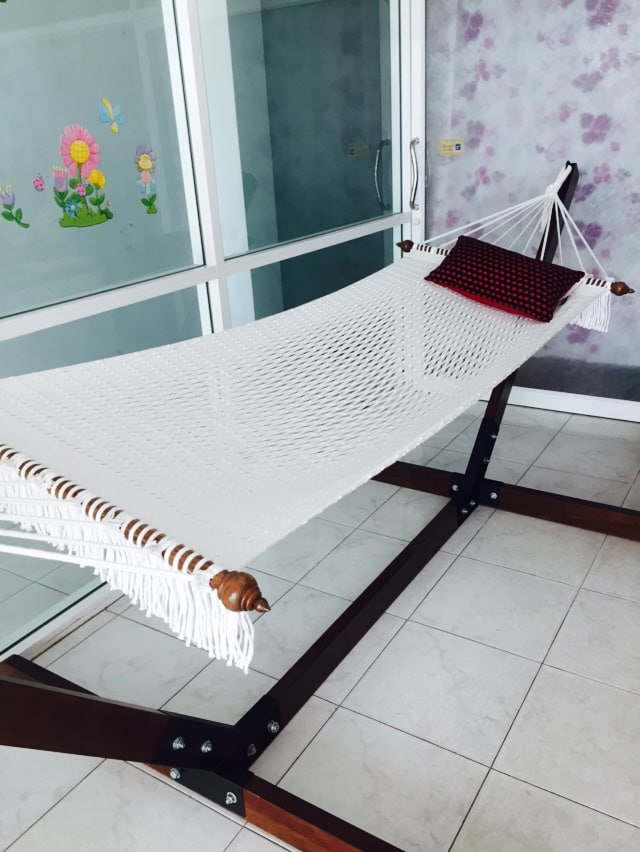 White cotton rope hammock with fringe, displayed on a dark wooden arc hammock stand on a tiled indoor patio with a small red cushion