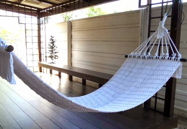 Woven white rope hammock with dark wood spreader bars hanging on an outdoor covered wooden deck or patio.