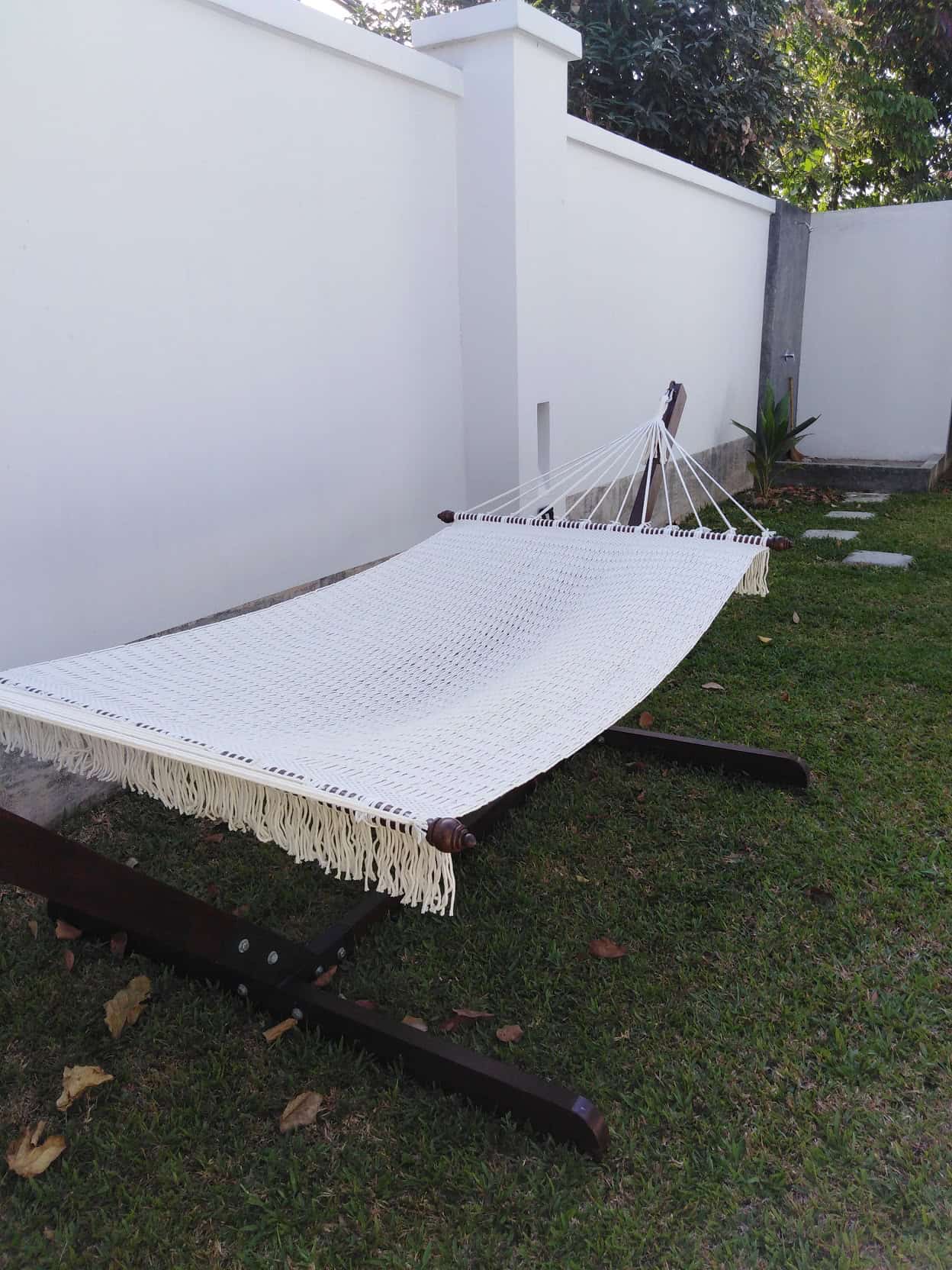 Outdoor beige cotton rope hammock and wooden stand on a grassy area next to a minimalist white wall.