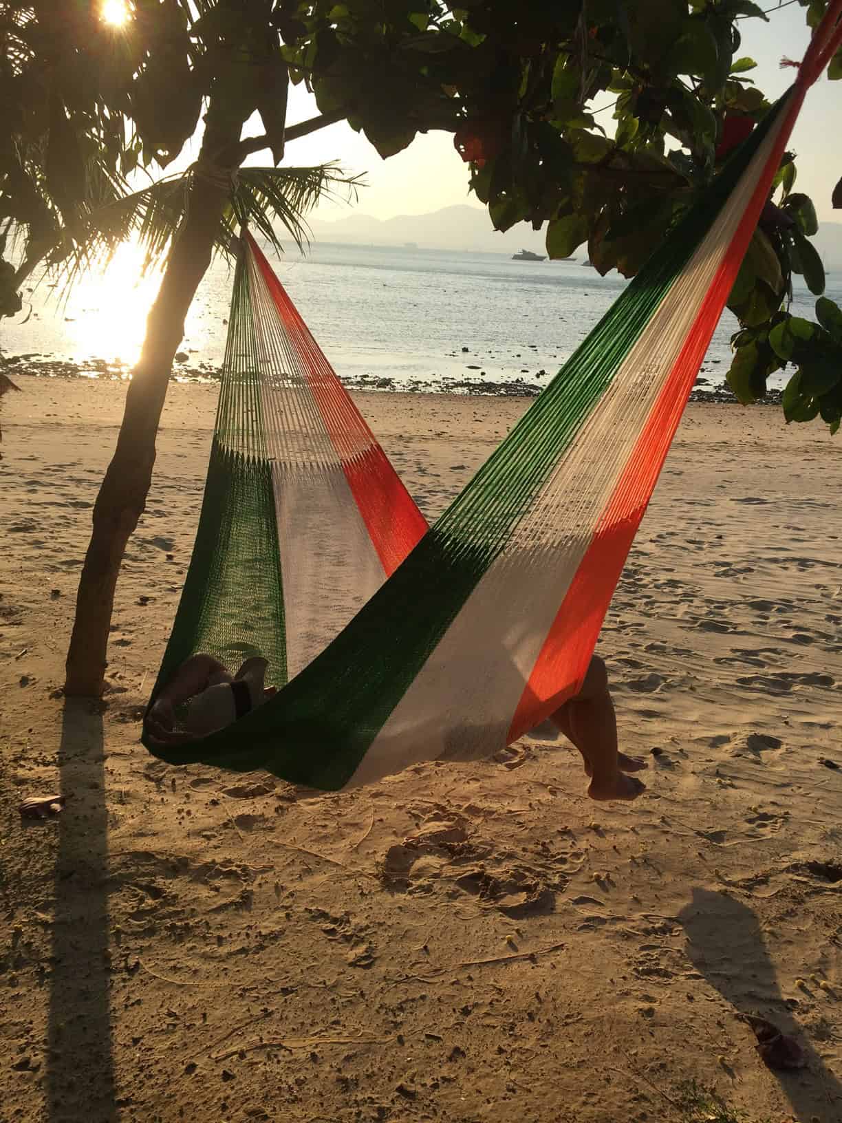 Green, white, and orange striped hammock hanging between two trees on a sandy beach during sunset, with a person relaxing inside.
