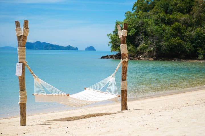 White rope hammock strung between two rustic wooden posts on a tropical sandy beach with clear turquoise ocean and islands in the background.
