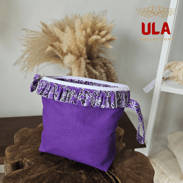 a square zippered pouch or clutch bag with a vibrant solid purple cotton body. The bag is accented with a ruffled trim along the white zipper, made from a black and white batik fabric featuring a floral or vine print. The bag also has a small wrist strap made from the same batik print fabric. This fashion accessory provides a bright pop of color and a bohemian or eclectic style, shown in a retail or closet setting.