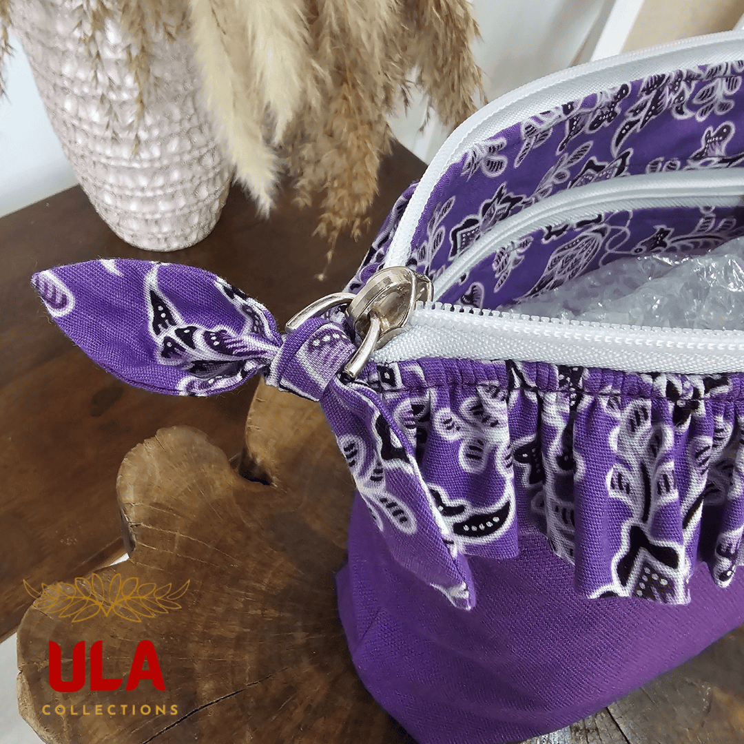 a square zippered pouch or clutch bag with a vibrant solid purple cotton body. The bag is accented with a ruffled trim along the white zipper, made from a black and white batik fabric featuring a floral or vine print. The bag also has a small wrist strap made from the same batik print fabric. This fashion accessory provides a bright pop of color and a bohemian or eclectic style, shown in a retail or closet setting.