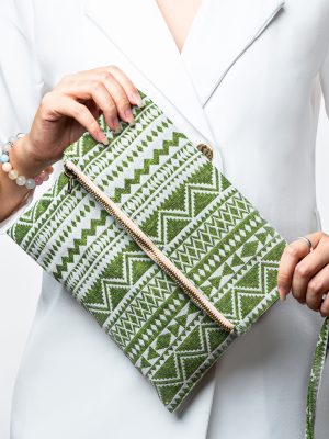 A stylish fold-over clutch bag or zippered wristlet made from a textured woven fabric featuring an ethnic or tribal geometric pattern. The textile is colored in various shades of light and bright green, forming repeating vertical stripes of chevron and diamond motifs. The clutch has a decorative trim along the zipper line and a matching patterned fabric wrist strap. The green accessory is displayed against a white background with dried grasses, promoting a bohemian or eclectic fashion style.