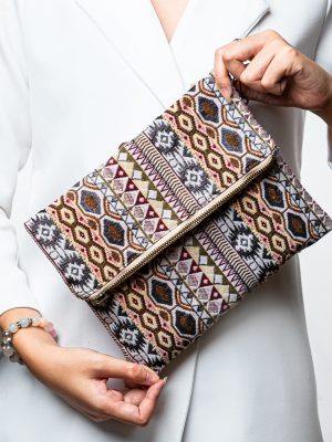 A stylish fold-over clutch bag or zippered wristlet made from a textured woven fabric featuring an intricate Southwestern or Aztec geometric pattern. The textile uses a rich, earthy color palette including shades of terracotta, dusty rose, brown, cream, and black. The clutch has a beige zigzag trim along the zipper line and a matching patterned fabric wrist strap. This bohemian fashion accessory is displayed on a white surface, ideal for everyday wear or travel.