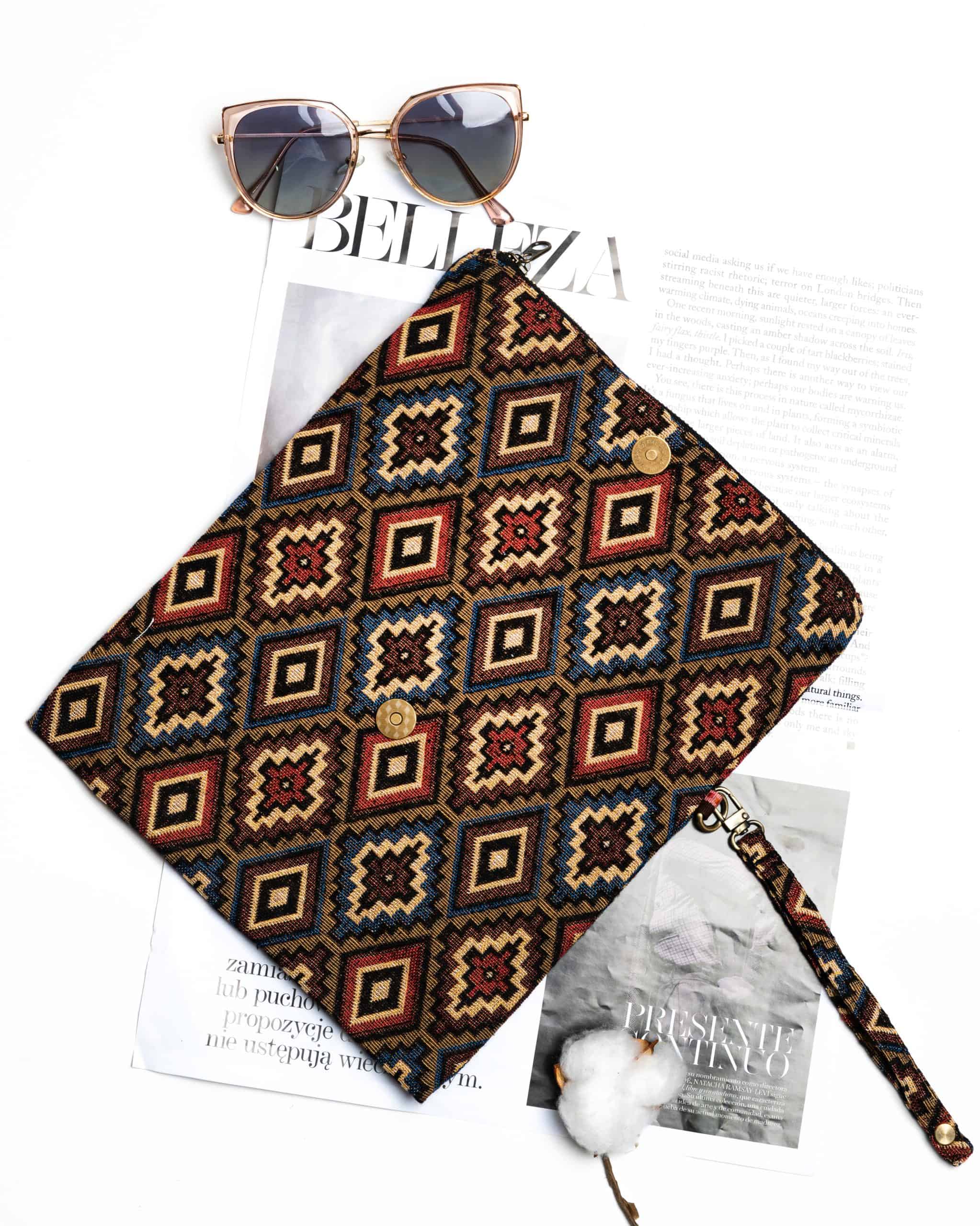 Here is the alt text for good SEO describing this image: A stylish fold-over clutch bag or zippered wristlet made from a dark, textured woven fabric. The bag features an intricate, repeating diamond geometric pattern in a rich, autumnal color palette including deep red, rust, light blue, and mustard yellow on a dark brown background. The clutch has a black zipper and a patterned fabric wrist strap. This bohemian fashion accessory is displayed on a magazine page, showcasing a maximalist or Southwestern style for everyday or travel use.