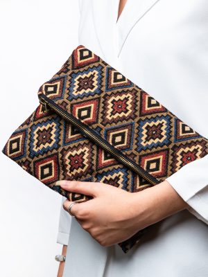 Here is the alt text for good SEO describing this image: A stylish fold-over clutch bag or zippered wristlet made from a dark, textured woven fabric. The bag features an intricate, repeating diamond geometric pattern in a rich, autumnal color palette including deep red, rust, light blue, and mustard yellow on a dark brown background. The clutch has a black zipper and a patterned fabric wrist strap. This bohemian fashion accessory is displayed on a magazine page, showcasing a maximalist or Southwestern style for everyday or travel use.