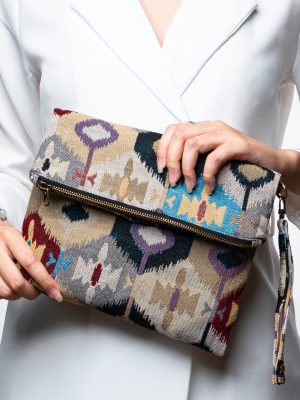 A stylish fold-over clutch bag or zippered wristlet made from a light-colored textured woven fabric. The bag features an intricate Southwestern or Ikat geometric pattern in muted, earthy tones including beige, gray, mustard yellow, dark teal, and burgundy. The clutch has a black zipper and a matching patterned fabric wrist strap. This bohemian fashion accessory is displayed on a magazine page, ideal for adding an eclectic tribal touch to an outfit for everyday wear or travel.