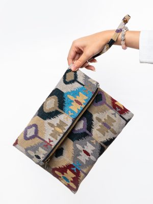 A stylish fold-over clutch bag or zippered wristlet made from a light-colored textured woven fabric. The bag features an intricate Southwestern or Ikat geometric pattern in muted, earthy tones including beige, gray, mustard yellow, dark teal, and burgundy. The clutch has a black zipper and a matching patterned fabric wrist strap. This bohemian fashion accessory is displayed on a magazine page, ideal for adding an eclectic tribal touch to an outfit for everyday wear or travel.