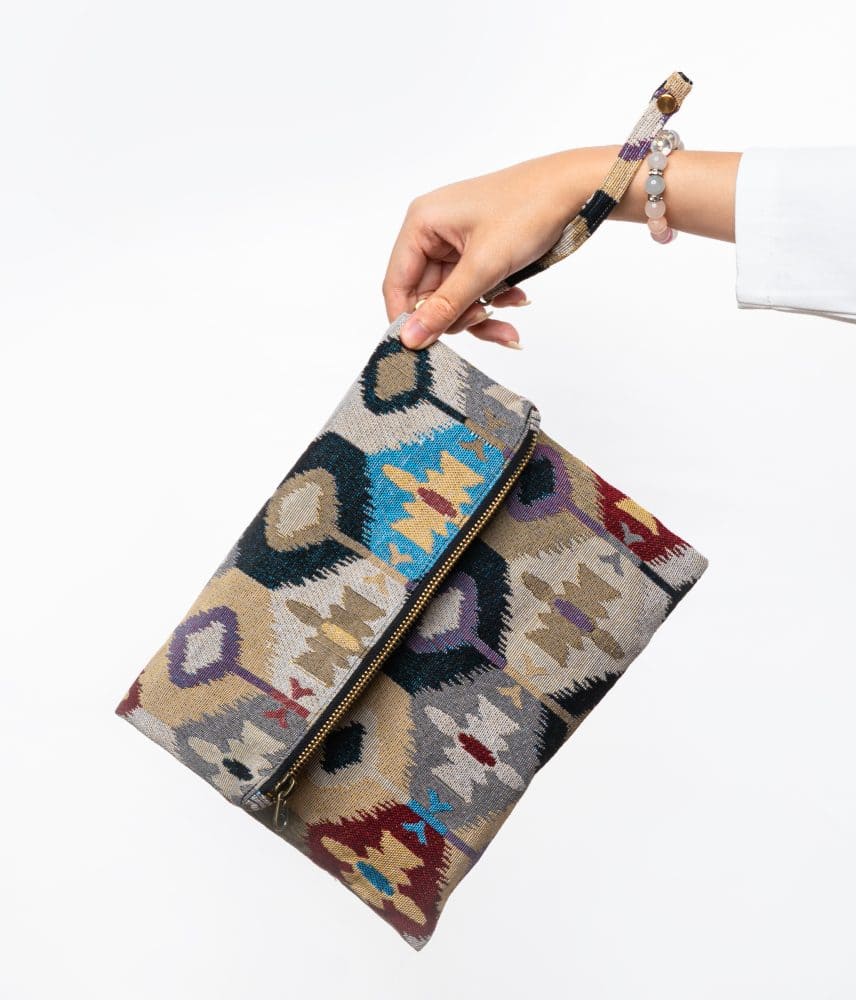 A stylish fold-over clutch bag or zippered wristlet made from a light-colored textured woven fabric. The bag features an intricate Southwestern or Ikat geometric pattern in muted, earthy tones including beige, gray, mustard yellow, dark teal, and burgundy. The clutch has a black zipper and a matching patterned fabric wrist strap. This bohemian fashion accessory is displayed on a magazine page, ideal for adding an eclectic tribal touch to an outfit for everyday wear or travel.
