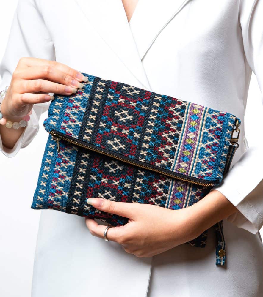 A stylish fold-over clutch bag or zippered wristlet made from a textured woven fabric featuring an ethnic or Aztec geometric pattern. The textile is primarily in shades of teal, deep blue, and cream, accented with dark burgundy, black, and gold woven details. The clutch has a metal zipper and a matching patterned fabric wrist strap, and is displayed against a white background, promoting it as a bohemian fashion accessory or travel handbag.
