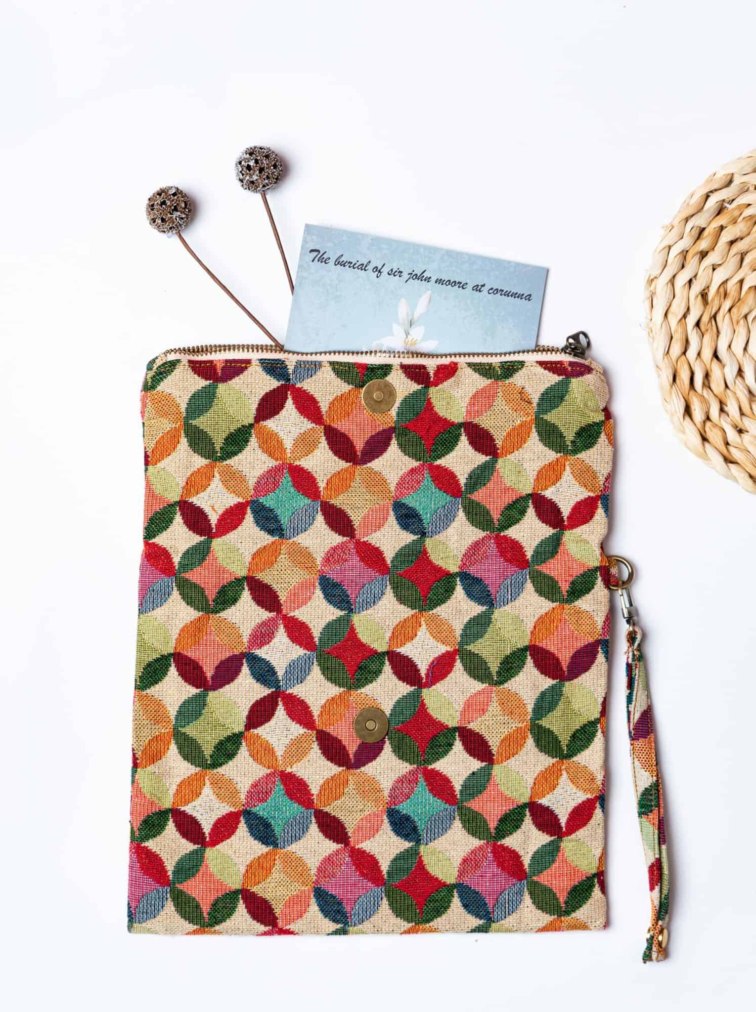 A stylish fold-over clutch bag or zippered wristlet made from a textured natural woven fabric. The bag features a vibrant, multi-color geometric pattern of interlocking circles or Seigaiha wave motifs in shades of red, teal, orange, green, and pink. The clutch has a white zipper and a matching patterned fabric wrist strap, and is displayed on a white surface with a cork coaster and wicker mat in the background, promoting it as a bohemian fashion accessory.