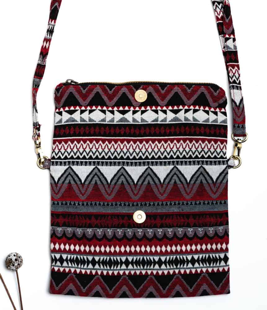 A stylish fold-over crossbody bag or clutch made from a textured woven fabric. The bag features an intricate tribal or geometric pattern with repeating chevron and diamond motifs in a high-contrast color palette of red, black, white, and gray. The accessory has a black zipper along the fold and a detachable, matching patterned fabric strap with gold-toned hardware, making it a versatile bohemian fashion accessory for everyday wear or travel.