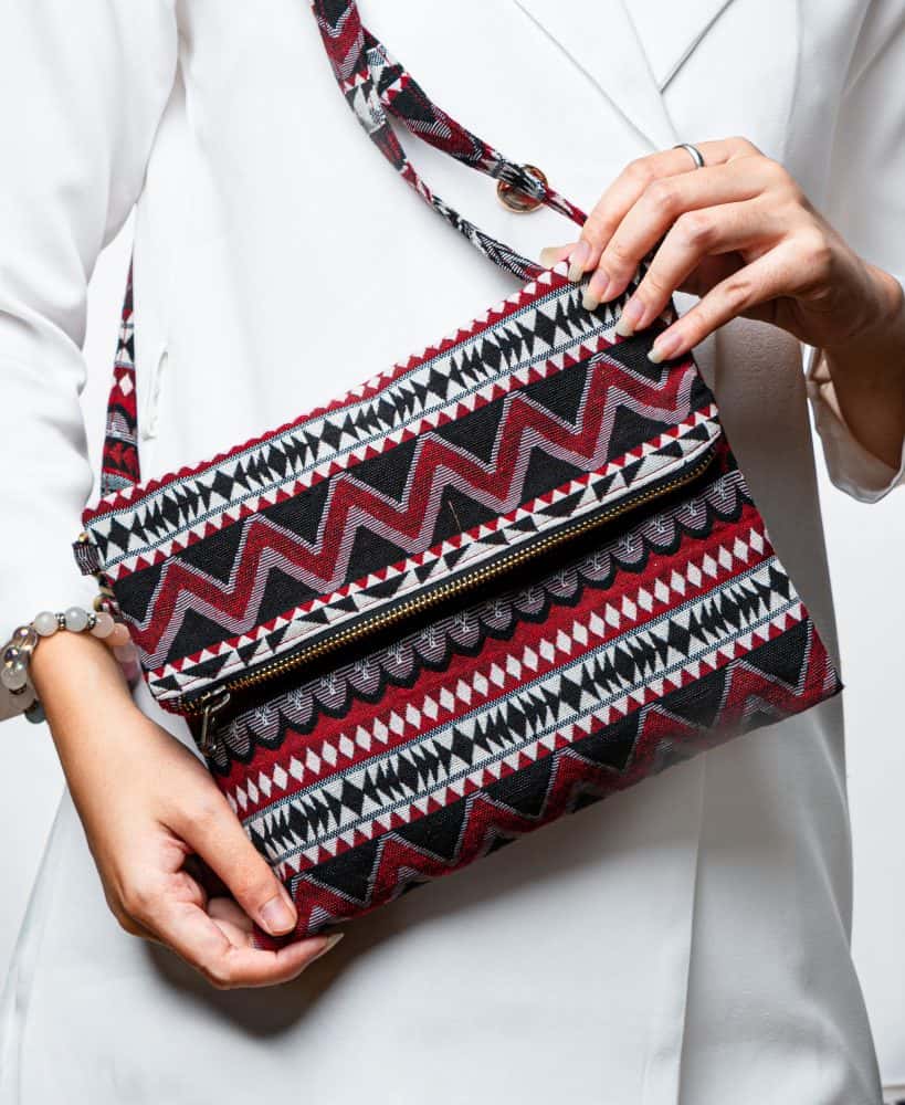 A stylish fold-over crossbody bag or clutch made from a textured woven fabric. The bag features an intricate tribal or geometric pattern with repeating chevron and diamond motifs in a high-contrast color palette of red, black, white, and gray. The accessory has a black zipper along the fold and a detachable, matching patterned fabric strap with gold-toned hardware, making it a versatile bohemian fashion accessory for everyday wear or travel.