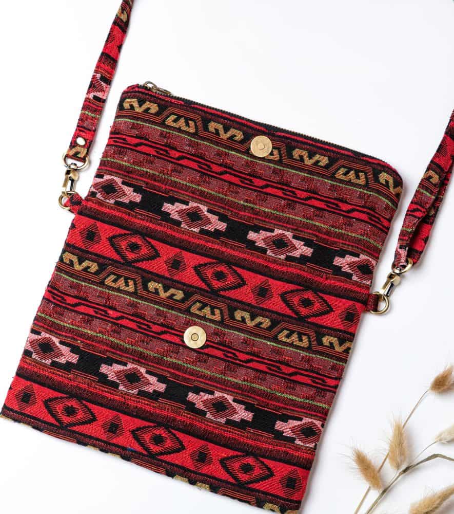 A person's hand is holding up a fold-over crossbody bag or shoulder bag made from a dark, textured woven fabric. The bag features an intricate tribal or geometric pattern with repeating motifs in a rich color palette of red, burgundy, black, and gold. The bag has a black zipper along the fold and a wide, detachable, matching patterned fabric strap with gold-toned hardware, showcasing a bohemian fashion accessory for everyday wear or travel.
