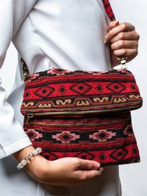 A person's hand is holding up a fold-over crossbody bag or shoulder bag made from a dark, textured woven fabric. The bag features an intricate tribal or geometric pattern with repeating motifs in a rich color palette of red, burgundy, black, and gold. The bag has a black zipper along the fold and a wide, detachable, matching patterned fabric strap with gold-toned hardware, showcasing a bohemian fashion accessory for everyday wear or travel.