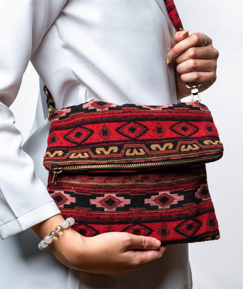 A person's hand is holding up a fold-over crossbody bag or shoulder bag made from a dark, textured woven fabric. The bag features an intricate tribal or geometric pattern with repeating motifs in a rich color palette of red, burgundy, black, and gold. The bag has a black zipper along the fold and a wide, detachable, matching patterned fabric strap with gold-toned hardware, showcasing a bohemian fashion accessory for everyday wear or travel.