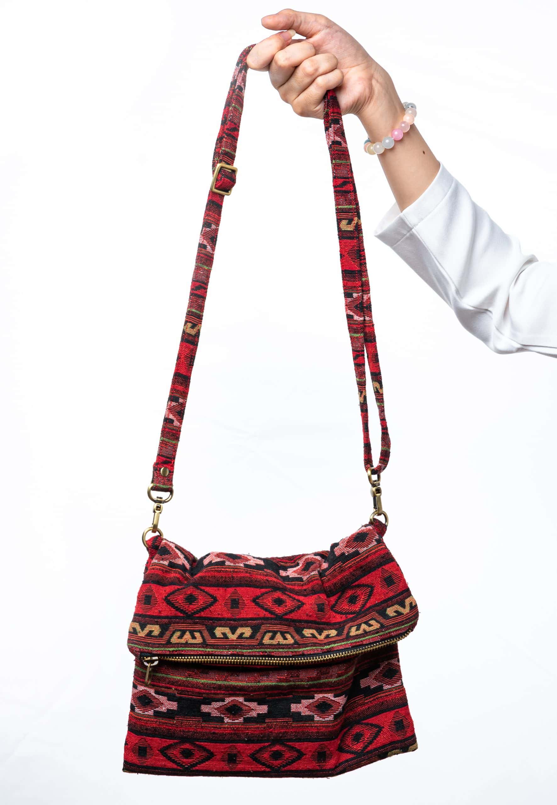 A person's hand is holding up a fold-over crossbody bag or shoulder bag made from a dark, textured woven fabric. The bag features an intricate tribal or geometric pattern with repeating motifs in a rich color palette of red, burgundy, black, and gold. The bag has a black zipper along the fold and a wide, detachable, matching patterned fabric strap with gold-toned hardware, showcasing a bohemian fashion accessory for everyday wear or travel.