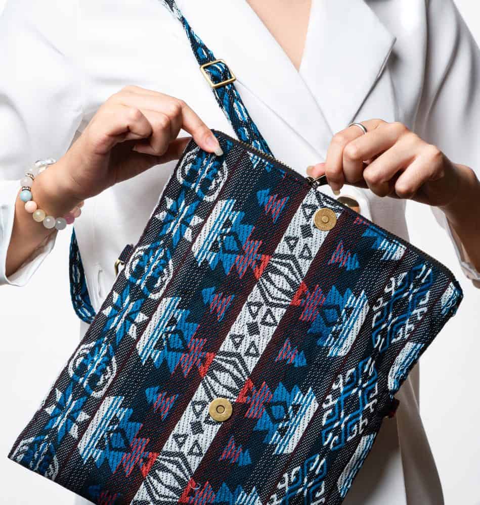 A stylish fold-over crossbody bag or shoulder bag made from a dark woven fabric. The bag features an intricate tribal or Ikat geometric pattern in a dramatic color palette of black, deep indigo, turquoise, dark red, and white. The bag has a zipper along the fold and a wide, matching patterned fabric strap with silver-toned hardware, showcasing a maximalist or eclectic fashion accessory for everyday wear or travel.