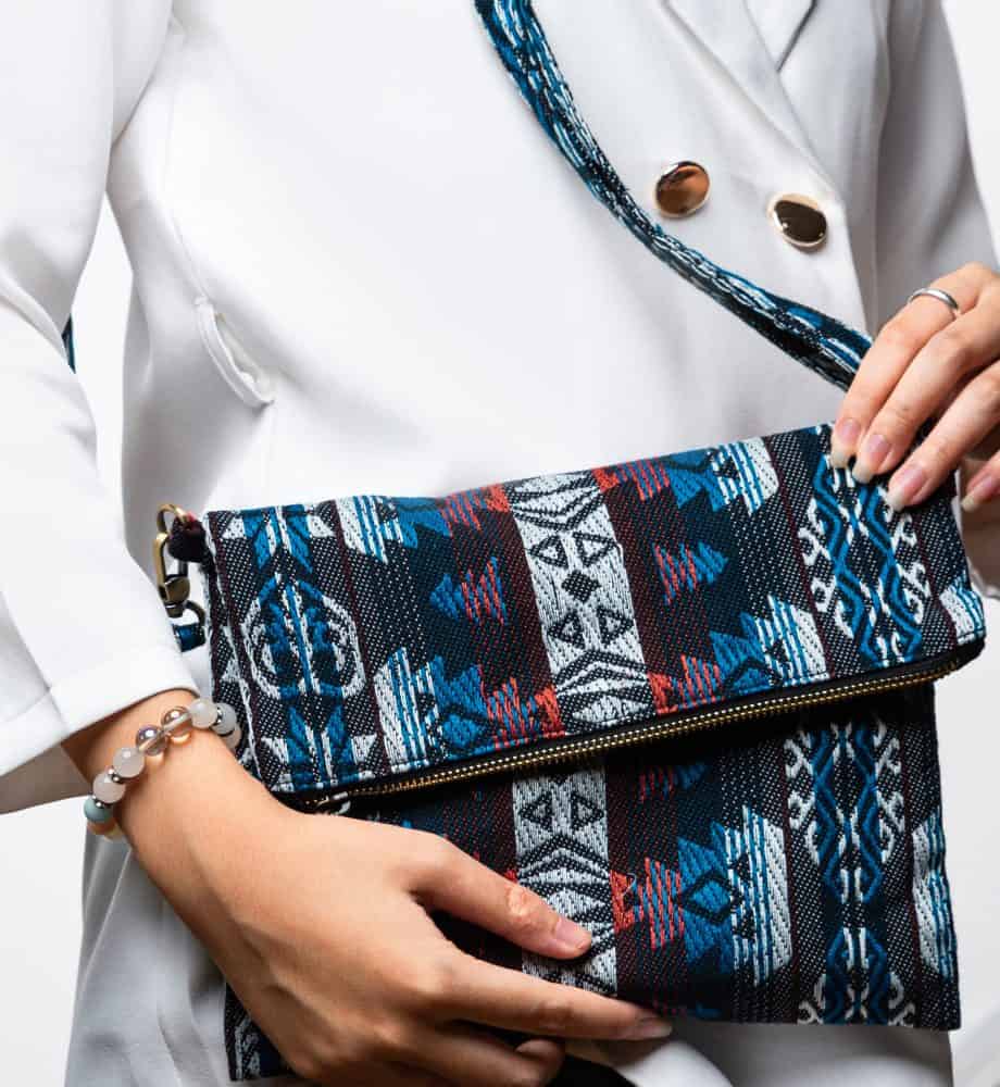 A stylish fold-over crossbody bag or shoulder bag made from a dark woven fabric. The bag features an intricate tribal or Ikat geometric pattern in a dramatic color palette of black, deep indigo, turquoise, dark red, and white. The bag has a zipper along the fold and a wide, matching patterned fabric strap with silver-toned hardware, showcasing a maximalist or eclectic fashion accessory for everyday wear or travel.