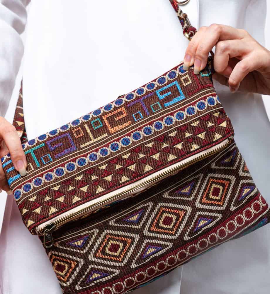 A stylish fold-over crossbody bag or shoulder bag made from a dark brown woven fabric. The bag features an intricate Southwestern or Aztec geometric pattern with horizontal stripes of diamond, zigzag, and Greek key motifs in shades of burgundy, deep blue, turquoise, and orange. The bag has a small zipper along the fold and a wide, matching patterned fabric strap with silver-toned hardware, showcasing an eclectic fashion accessory for everyday wear or travel.