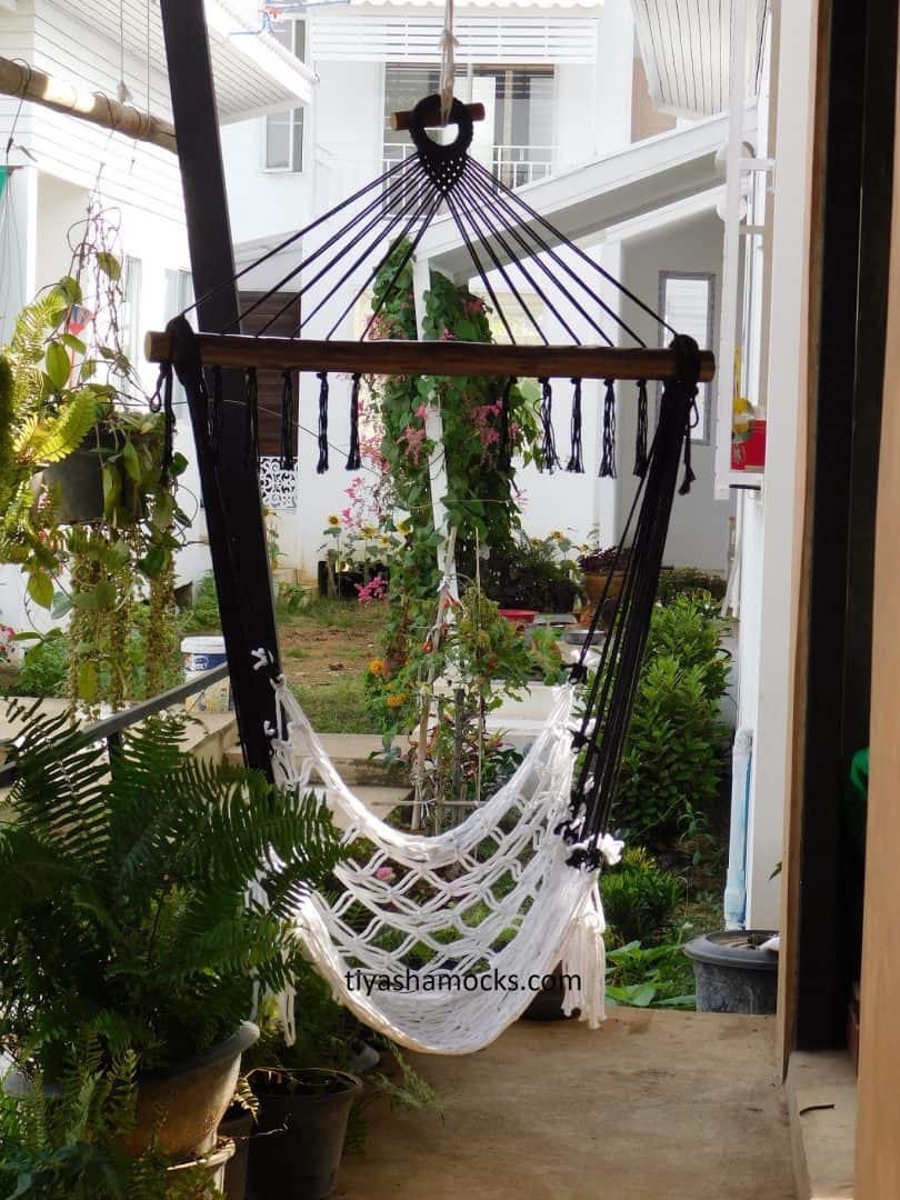Pausa hammock chair with black hanging ropes and white woven net, set up on a porch with ferns and climbing plants in a bright outdoor residential environment.