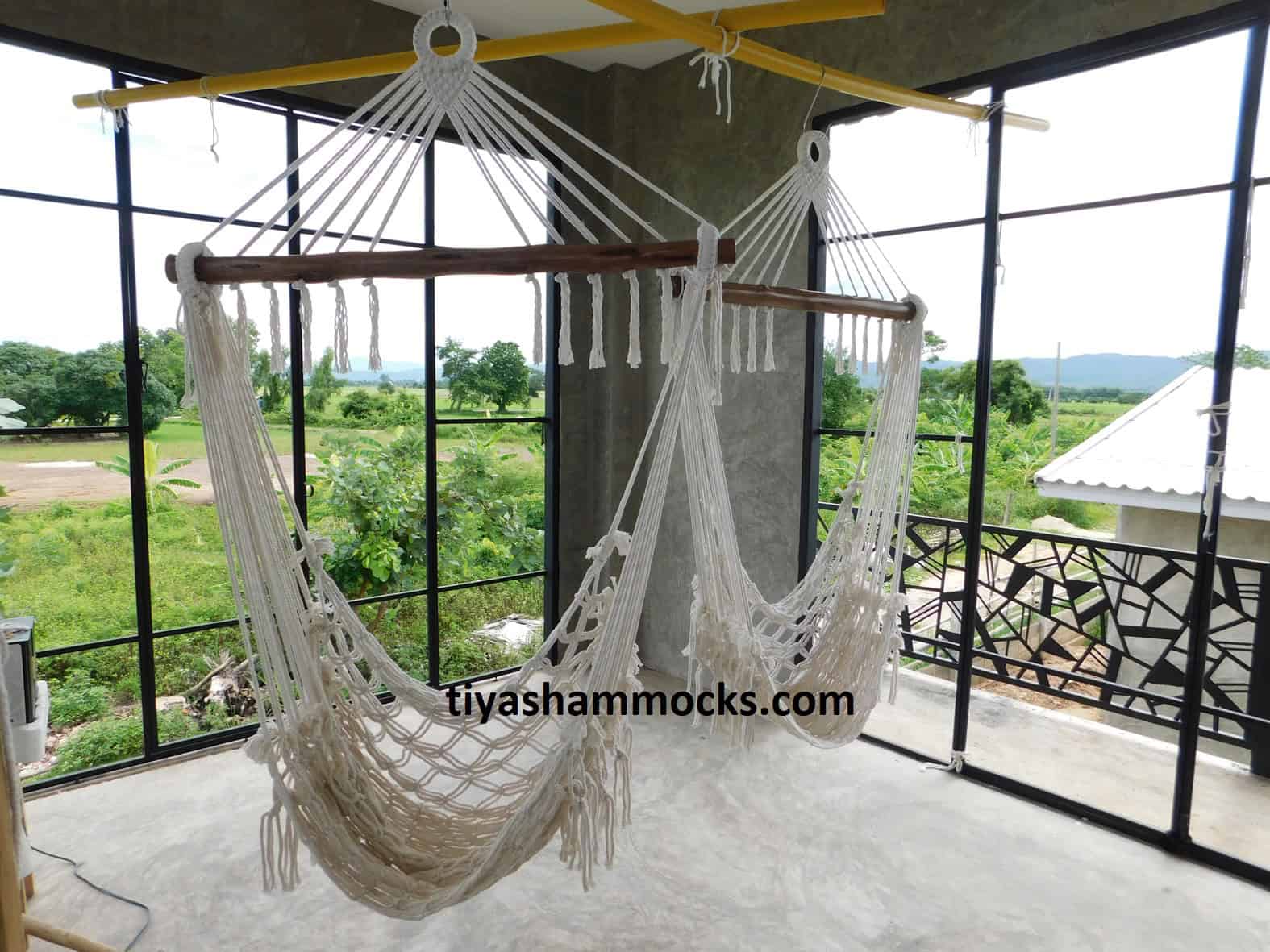 Two natural beige macrame "Pausa" hammock chairs with wooden bars, hung on a stylish balcony overlooking a lush green field and countryside environment.