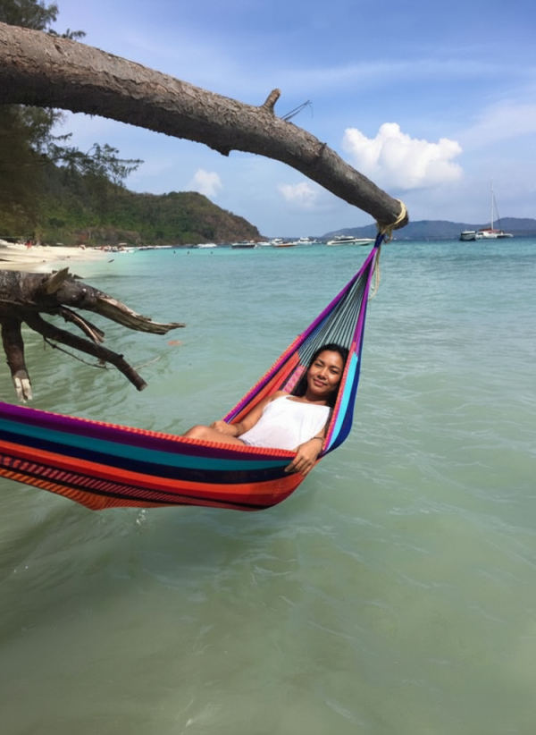 Woman smiling in a vibrant striped hammock suspended over clear turquoise ocean water on a sunny beach.