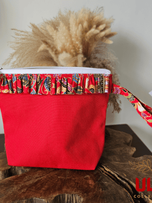 A vibrant zippered pouch bag or clutch with a solid red cotton body. The bag is decorated with a wide, ruffled trim and interior lining made from a red batik fabric with a multicolor floral print in shades of gold, teal, and brown. The bag is shown open and includes a matching batik fabric wrist strap or pull tab. It is resting on a dark, rustic wooden surface, with a logo and dried pampas grass in the background, highlighting an eclectic fashion accessory perfect for travel or a statement handbag.