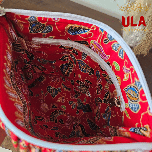 A vibrant zippered pouch bag or clutch with a solid red cotton body. The bag is decorated with a wide, ruffled trim and interior lining made from a red batik fabric with a multicolor floral print in shades of gold, teal, and brown. The bag is shown open and includes a matching batik fabric wrist strap or pull tab. It is resting on a dark, rustic wooden surface, with a logo and dried pampas grass in the background, highlighting an eclectic fashion accessory perfect for travel or a statement handbag.