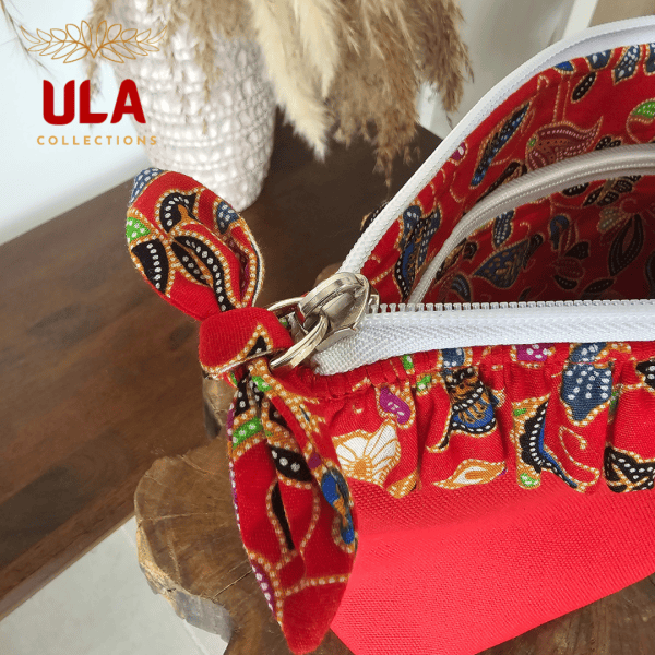 A vibrant zippered pouch bag or clutch with a solid red cotton body. The bag is decorated with a wide, ruffled trim and interior lining made from a red batik fabric with a multicolor floral print in shades of gold, teal, and brown. The bag is shown open and includes a matching batik fabric wrist strap or pull tab. It is resting on a dark, rustic wooden surface, with a logo and dried pampas grass in the background, highlighting an eclectic fashion accessory perfect for travel or a statement handbag.