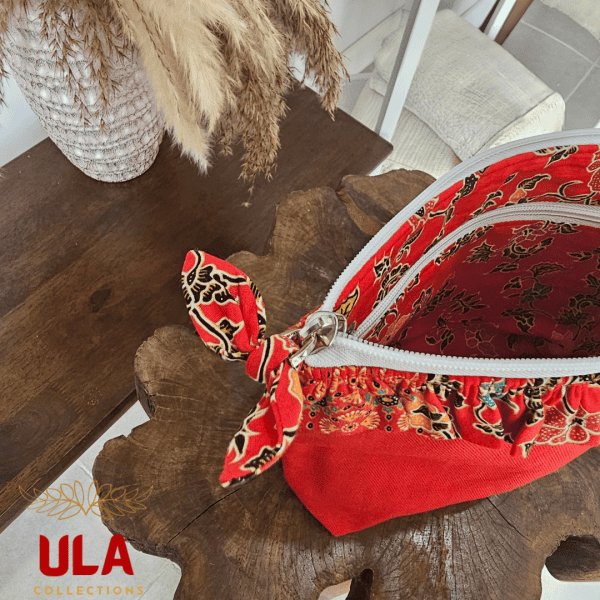 A vibrant zippered pouch bag or clutch with a solid red cotton body. The bag is decorated with a wide, ruffled trim and interior lining made from a red batik fabric with a multicolor floral print in shades of gold, teal, and brown. The bag is shown open and includes a matching batik fabric wrist strap or pull tab. It is resting on a dark, rustic wooden surface, with a logo and dried pampas grass in the background, highlighting an eclectic fashion accessory perfect for travel or a statement handbag.
