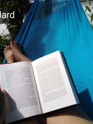 A close-up view of a person relaxing and reading a book while lying in a bright blue woven hammock outdoors. The shot focuses on the reader's hands and bare feet, with lush green tropical foliage and a white wall visible in the background, promoting a feeling of travel, relaxation, and outdoor leisure.