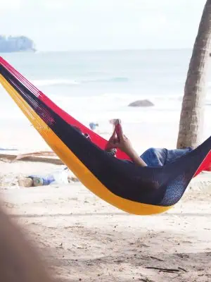 Person relaxing and reading a phone in a red, black, and yellow striped hammock tied between palm trees on a sunny tropical beach with the ocean in the background.