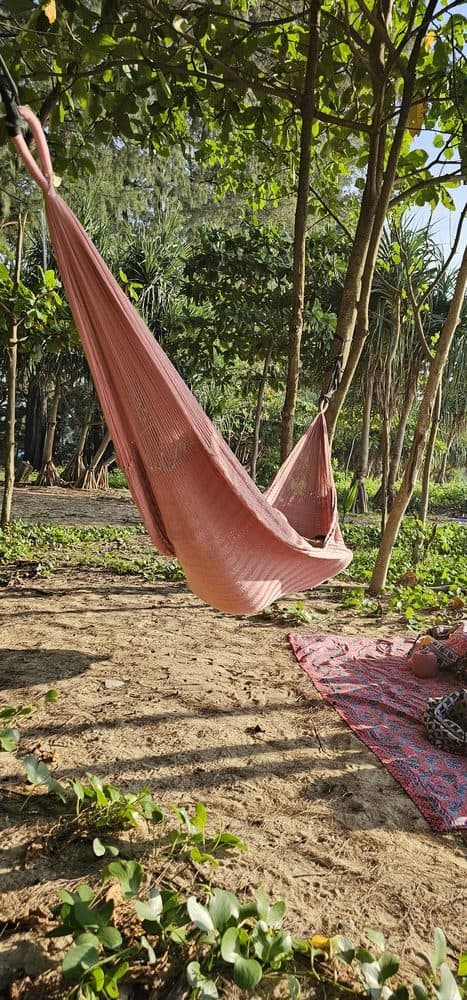 ULA "Copper Rose" hammock in a solid burnt orange/copper tone, hanging between trees on a tropical beach with sand and lush foliage.