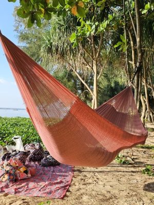 ULA "Copper Rose" hammock in a solid burnt orange/copper tone, hanging between trees on a tropical beach with sand and lush foliage.