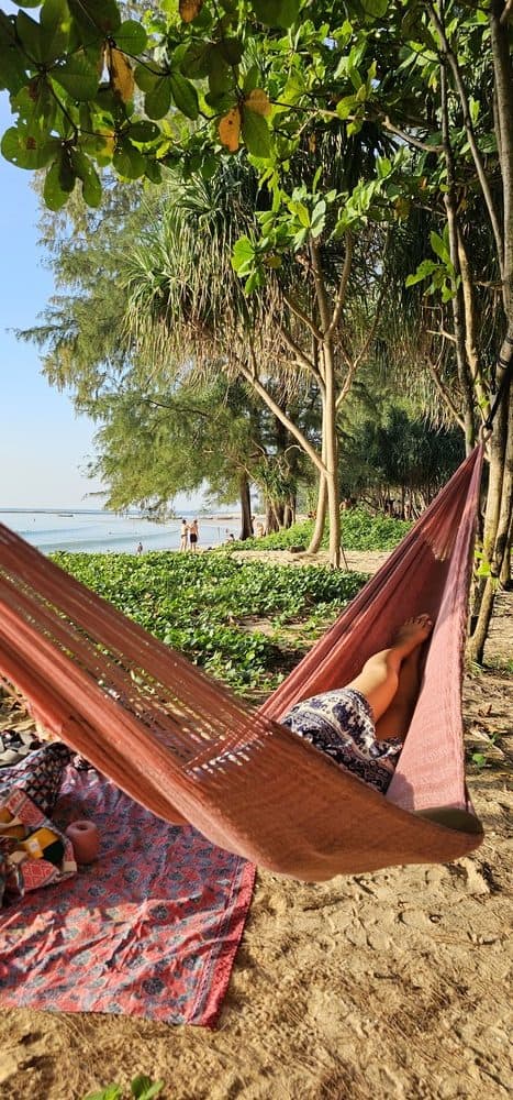ULA "Copper Rose" hammock in a solid burnt orange/copper tone, hanging between trees on a tropical beach with sand and lush foliage.