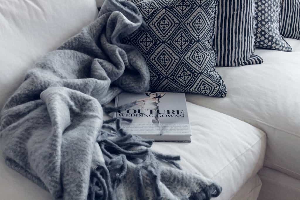 A stylish living room featuring a white sofa with patterned pillows, a blanket, and a fashion book.