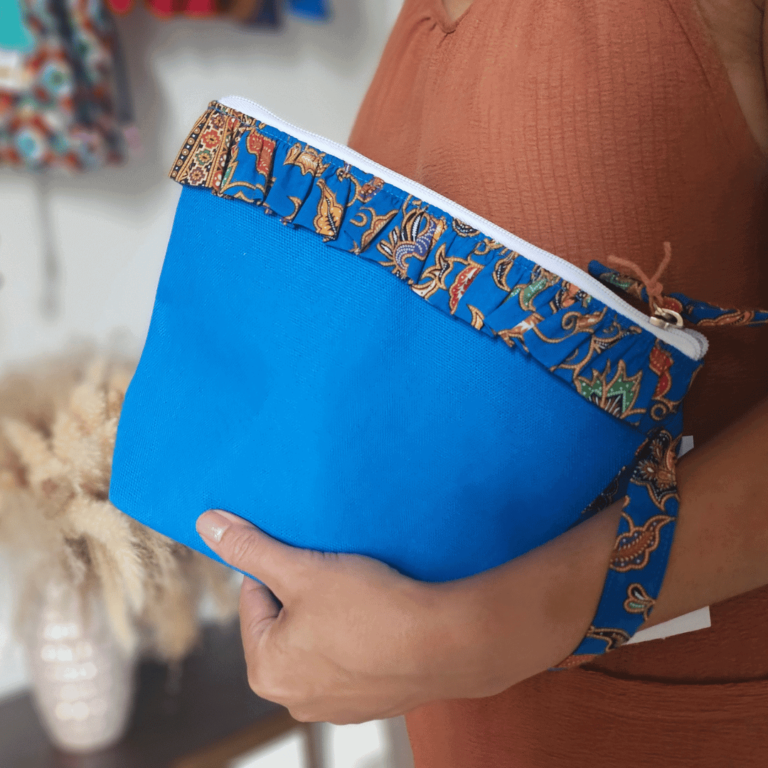 A vibrant zippered pouch bag or clutch with a solid royal blue cotton body. The bag is decorated with a wide, ruffled trim along the white zipper, made from a blue batik fabric with a multicolor floral print in shades of gold, purple, and red. The bag is shown open and includes a matching batik fabric wrist strap. It rests on a dark, rustic wooden surface, with a logo and dried pampas grass in the background, showcasing an eclectic fashion accessory perfect for travel or casual use.