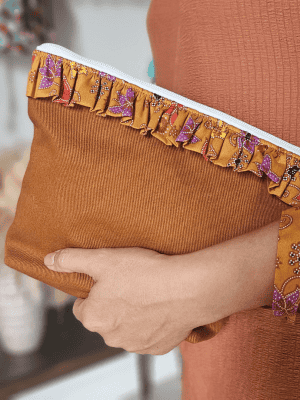A stylish zippered pouch or clutch bag featuring a deep ochre brown cotton body with a subtle texture. The top is decorated with a wide, ruffled trim and interior lining made from a vibrant gold batik fabric with purple and brown floral motifs. This versatile fashion accessory includes a matching batik wrist strap and is shown styled as a handbag and resting on a wooden surface, offering an eclectic and bohemian accent for everyday or travel use.