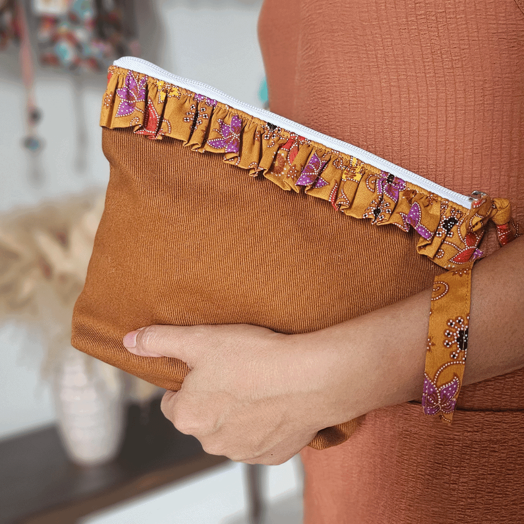 A stylish zippered pouch or clutch bag featuring a deep ochre brown cotton body with a subtle texture. The top is decorated with a wide, ruffled trim and interior lining made from a vibrant gold batik fabric with purple and brown floral motifs. This versatile fashion accessory includes a matching batik wrist strap and is shown styled as a handbag and resting on a wooden surface, offering an eclectic and bohemian accent for everyday or travel use.