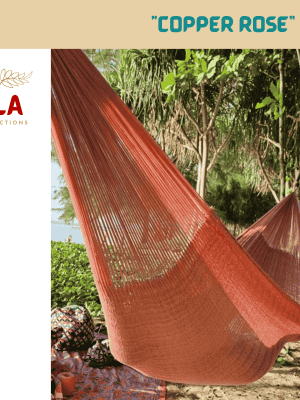 ULA "Copper Rose" hammock in a solid burnt orange/copper tone, hanging between trees on a tropical beach with sand and lush foliage.