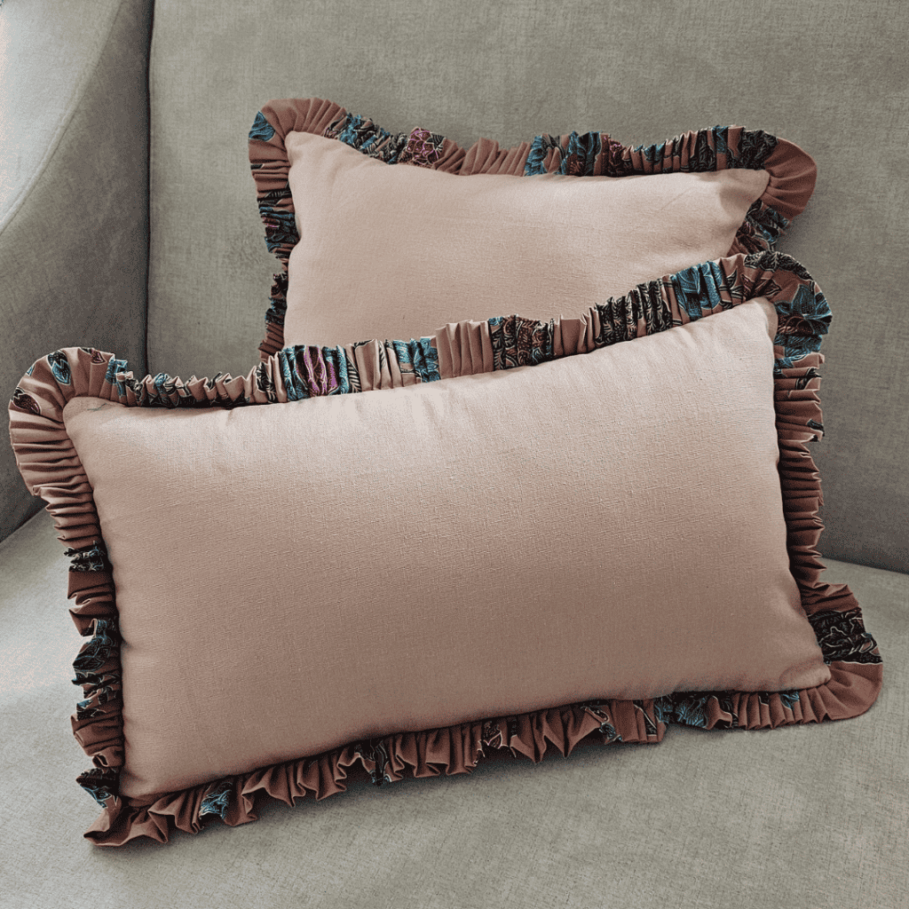 Two rectangular lumbar throw pillows with a dusky pink blush cotton body and a wide, ruffled trim featuring a batik cotton floral fabric edge. The batik fabric displays a vibrant, multicolor pattern in shades of teal, purple, fuchsia, and navy. The decorative cushions are resting on a neutral gray sofa, blending soft cotton texture and eclectic color for stylish home decor and interior design emphasis.