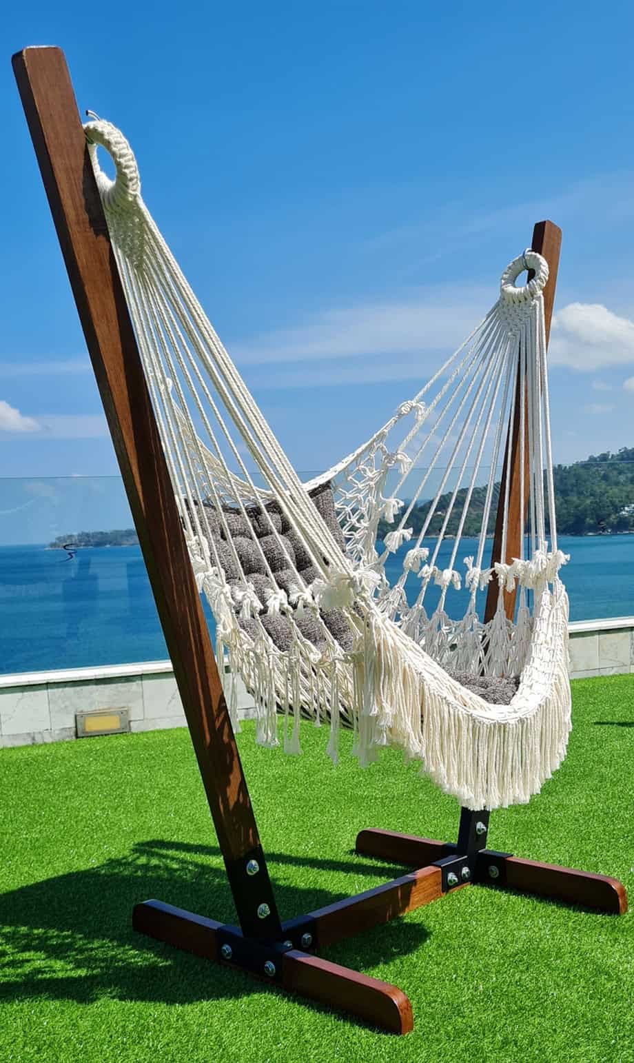 White macrame hammock chair with decorative fringe, hanging on a dark wood A-frame stand. Set up outdoors on a bright green turf with a stunning tropical ocean view in the background.