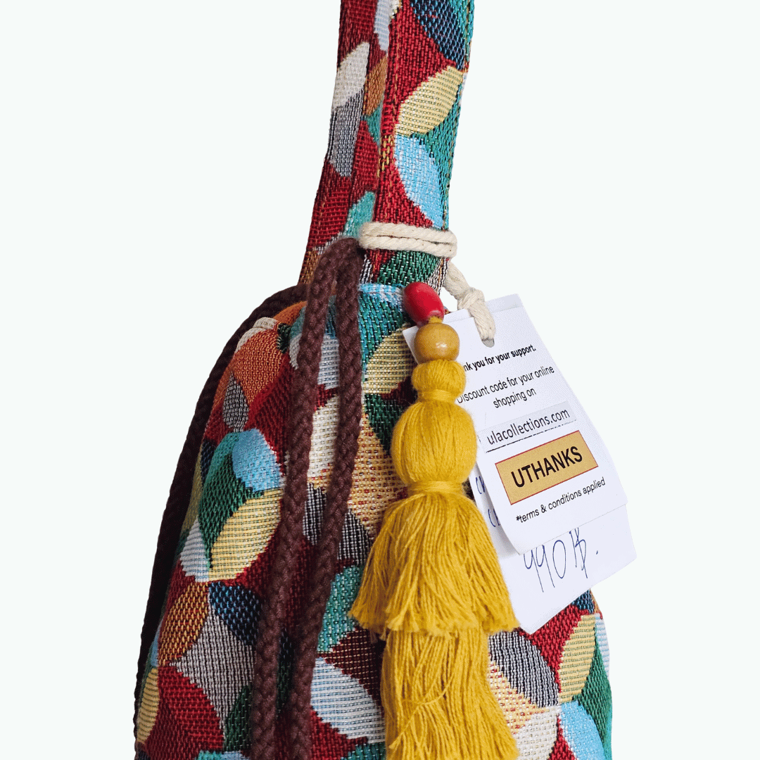 A stylish drawstring bucket bag or pouch bag featuring a vibrant, multi-color geometric fabric pattern with interlocking circle or Seigaiha wave motifs with bohemian style. The textile includes shades of teal, burgundy, olive green, cream, and brown. Decorated with tassels and wood beats