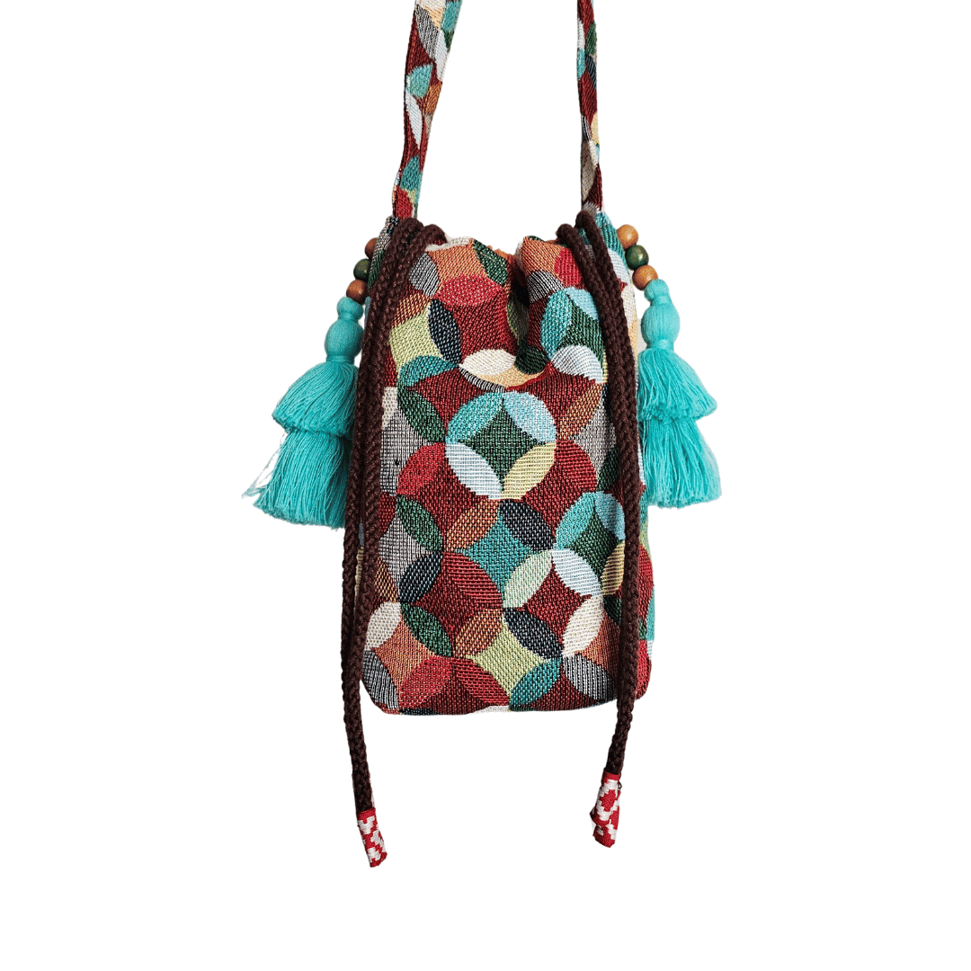 A stylish drawstring bucket bag or pouch bag featuring a vibrant, multi-color geometric fabric pattern with interlocking circle or Seigaiha wave motifs with bohemian style. The textile includes shades of teal, burgundy, olive green, cream, and brown. Decorated with tassels and wood beats